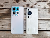 Two smartphones are placed side by side on a wooden surface. The phone on the left is a model from Infinix with a silver back and multiple camera lenses, one of which features a blue and pink-hued casing. The phone on the right is a model from Huawei, featuring a white back with a textured pattern and a prominent black camera module.