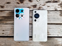 Two smartphones are placed side by side on a wooden surface. The phone on the left is a model from Infinix with a silver back and multiple camera lenses, one of which features a blue and pink-hued casing. The phone on the right is a model from Huawei, featuring a white back with a textured pattern and a prominent black camera module.