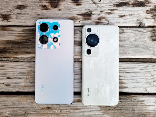 Two smartphones are placed side by side on a wooden surface. The phone on the left is a model from Infinix with a silver back and multiple camera lenses, one of which features a blue and pink-hued casing. The phone on the right is a model from Huawei, featuring a white back with a textured pattern and a prominent black camera module.
