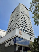 A modern high-rise building with an innovative facade design, featuring staggered windows creating a geometric pattern. The structure is set against a clear blue sky, with greenery visible to the side, adding a natural element. The lower part of the building showcases reflective glass surfaces.