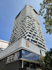 A modern high-rise building with an innovative facade design, featuring staggered windows creating a geometric pattern. The structure is set against a clear blue sky, with greenery visible to the side, adding a natural element. The lower part of the building showcases reflective glass surfaces.