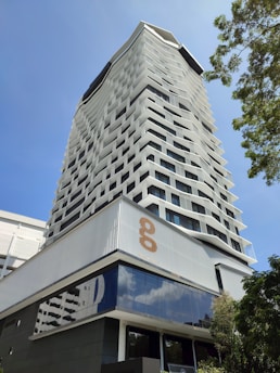 A modern high-rise building with an innovative facade design, featuring staggered windows creating a geometric pattern. The structure is set against a clear blue sky, with greenery visible to the side, adding a natural element. The lower part of the building showcases reflective glass surfaces.