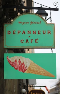 A green shop sign with a vintage design, featuring the text 'Magasin général Dépanneur Café' in red and a lower panel illustrating a stylized ice cream cone with red and white swirls.