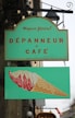 A green shop sign with a vintage design, featuring the text 'Magasin g&eacute;n&eacute;ral D&eacute;panneur Caf&eacute;' in red and a lower panel illustrating a stylized ice cream cone with red and white swirls.