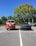 A bright red vintage Volkswagen van and a modern gray sedan are parked next to each other in a parking lot under a clear blue sky. The area is surrounded by lush green trees, and each vehicle is parked within designated parking spaces.