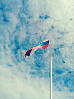 A Russian flag is waving on a tall flagpole against a background of a blue sky filled with scattered clouds. The colors of the flag, white, blue, and red, are clearly visible.