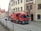 A red Coca-Cola delivery truck is parked on a cobblestone street in a European town. The truck is positioned near traditional, multi-story buildings. People and other vehicles are seen in the background, adding to the bustling urban scene.