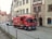 A red Coca-Cola delivery truck is parked on a cobblestone street in a European town. The truck is positioned near traditional, multi-story buildings. People and other vehicles are seen in the background, adding to the bustling urban scene.