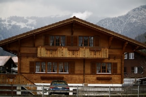 A traditional wooden chalet with intricate detailing, featuring a central balcony and multiple windows with shutters. Snow-capped mountains and overcast skies provide a picturesque backdrop. A car is parked in front of the house, and a wooden fence borders the property.