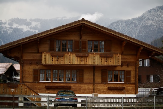 A traditional wooden chalet with intricate detailing, featuring a central balcony and multiple windows with shutters. Snow-capped mountains and overcast skies provide a picturesque backdrop. A car is parked in front of the house, and a wooden fence borders the property.