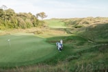 A lush, green golf course stretches across the landscape with several people gathered near the edge of one of the fairways. The scene is surrounded by gentle hills and dense clusters of trees under a clear sky.