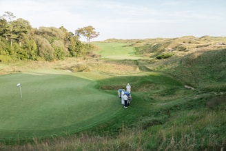 A lush, green golf course stretches across the landscape with several people gathered near the edge of one of the fairways. The scene is surrounded by gentle hills and dense clusters of trees under a clear sky.