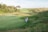 A lush, green golf course stretches across the landscape with several people gathered near the edge of one of the fairways. The scene is surrounded by gentle hills and dense clusters of trees under a clear sky.