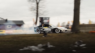 A race car with the branding 'Baström Motorsport' is drifting, producing smoke. It is outdoors with trees, a building in the background, and an overcast sky.