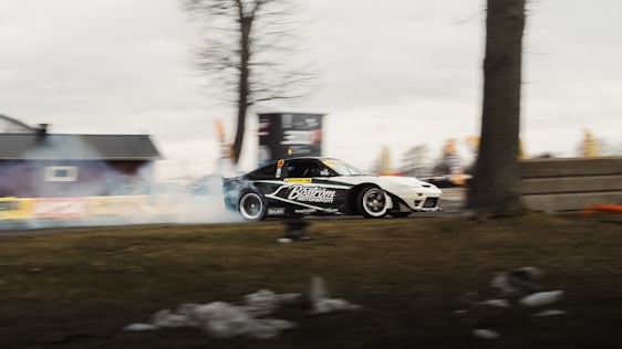 A race car with the branding 'Baström Motorsport' is drifting, producing smoke. It is outdoors with trees, a building in the background, and an overcast sky.