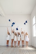 A group of six young women in white dresses celebrate by tossing blue graduation caps into the air inside a spacious room with white brick walls. The floor is wooden and there is natural light coming through windows on the right side.