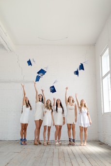 A group of six young women in white dresses celebrate by tossing blue graduation caps into the air inside a spacious room with white brick walls. The floor is wooden and there is natural light coming through windows on the right side.