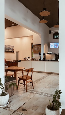 A cozy, modern cafe interior with wooden furniture, including a round table and chairs. White walls and a tiled floor add to the clean, minimalistic look. Hanging lights and a small corner counter with a coffee machine create a welcoming atmosphere. Potted plants add a touch of greenery.