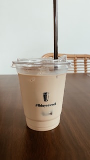 A plastic cup filled with iced coffee or a similar beverage sits on a wooden table. The cup features a lid with a straw and has '#8daysaweek' printed along with a small illustration of a glass. The background includes a wicker chair and a plain wall, giving a cozy and minimalist vibe.
