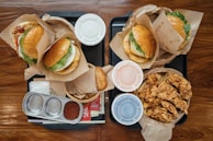 An overhead view of a meal consisting of several hamburgers wrapped in paper, a bowl of fried chicken pieces, small containers of dipping sauce, and a hash brown patty on a tray. The food is placed on a wooden table, and there are also metal and plastic containers present.