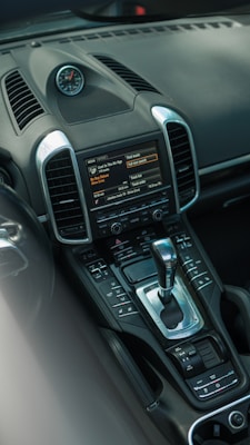 The image features the interior of a car showcasing a close-up of the central console. The console includes a sleek gear shift lever, various control buttons, and a touch screen display showing media options. There is also a clock mounted on top of the dashboard, adding a touch of elegance. The materials appear to be a combination of polished metal and dark leather or plastic, giving a luxurious and modern feel.