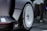 A close-up view of a futuristic car wheel with a sleek, aerodynamic design. The wheel features a white circular hubcap with the Hyundai logo at its center. The surrounding area of the car is shiny and metallic, with sharp angles and a minimalist aesthetic. In the background, an out-of-focus figure stands on a dark tiled floor, adding depth to the image.