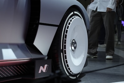 A close-up view of a futuristic car wheel with a sleek, aerodynamic design. The wheel features a white circular hubcap with the Hyundai logo at its center. The surrounding area of the car is shiny and metallic, with sharp angles and a minimalist aesthetic. In the background, an out-of-focus figure stands on a dark tiled floor, adding depth to the image.