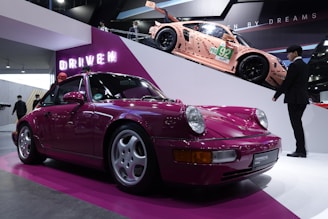 A sleek, magenta-colored classic Porsche is prominently displayed on a low pedestal with neon signage in the background reading 'DRIVEN'. Above, on a ramp, a modern racing car in peach with green and black detailing is visible, creating a dynamic contrast. Several people are around, including a man in a suit, seemingly admiring the vehicles.