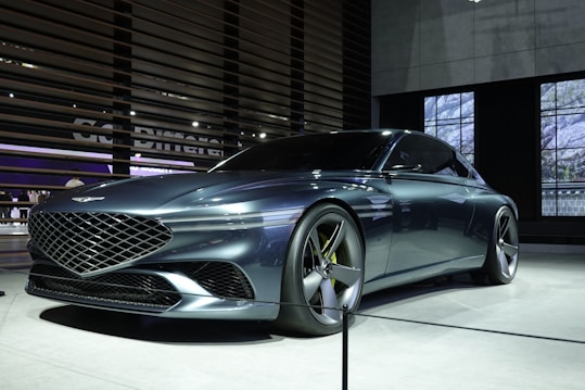 A sleek, futuristic car with a glossy metallic finish, showcased indoors. The vehicle features a large, intricate grille and smooth aerodynamic lines. It is displayed on a well-lit platform with spotlights accentuating its design.