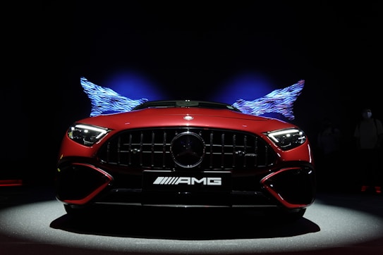 A sleek red sports car is prominently displayed against a dark background. The car's grill and headlights are aggressively designed, and it features the AMG logo at the front. Illuminated patterns resembling wings are positioned symmetrically behind the car, casting a dynamic and futuristic effect.