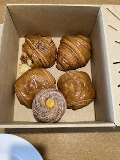 A cardboard box contains four pastries, including three croissants and one pastry dusted with sugar and topped with a dollop of yellow cream. The croissants have a glossy, golden-brown appearance, and the box is placed on a wooden surface.