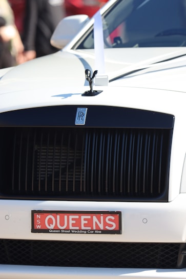 The front of a luxury car featuring a prominent grille and a small statue on the hood. A decorative white ribbon is attached to the hood, signifying that the vehicle is likely used for a wedding. The license plate reads 'QUEENS' and indicates a wedding car hire service.