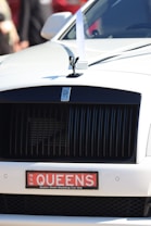 The front of a luxury car featuring a prominent grille and a small statue on the hood. A decorative white ribbon is attached to the hood, signifying that the vehicle is likely used for a wedding. The license plate reads 'QUEENS' and indicates a wedding car hire service.