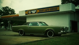 A vintage car with a muted green color is parked in front of an old-style auto service center. The building has a sign for 'BRAD'S AUTO SERVICE' and branding for 'NAPA AUTOCARE CENTER'. The location appears somewhat deserted, with an overcast sky and a few trees in the background.