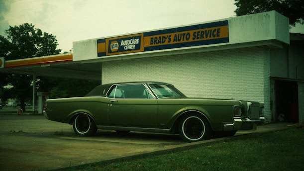 A vintage car with a muted green color is parked in front of an old-style auto service center. The building has a sign for 'BRAD'S AUTO SERVICE' and branding for 'NAPA AUTOCARE CENTER'. The location appears somewhat deserted, with an overcast sky and a few trees in the background.