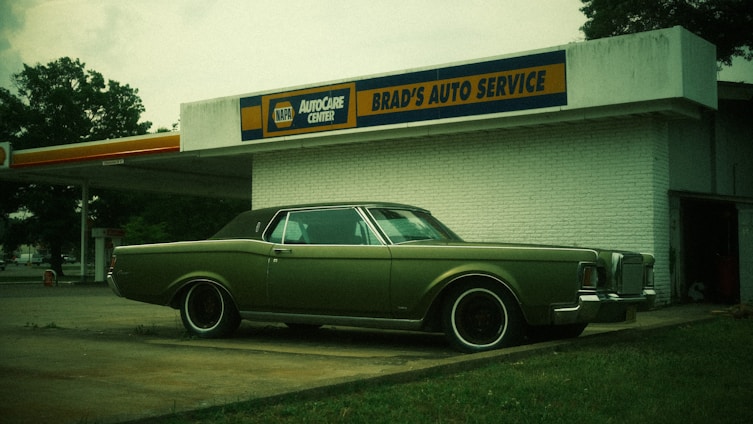 A vintage car with a muted green color is parked in front of an old-style auto service center. The building has a sign for 'BRAD'S AUTO SERVICE' and branding for 'NAPA AUTOCARE CENTER'. The location appears somewhat deserted, with an overcast sky and a few trees in the background.