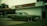 A vintage car with a muted green color is parked in front of an old-style auto service center. The building has a sign for 'BRAD'S AUTO SERVICE' and branding for 'NAPA AUTOCARE CENTER'. The location appears somewhat deserted, with an overcast sky and a few trees in the background.