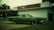 A vintage car with a muted green color is parked in front of an old-style auto service center. The building has a sign for 'BRAD'S AUTO SERVICE' and branding for 'NAPA AUTOCARE CENTER'. The location appears somewhat deserted, with an overcast sky and a few trees in the background.