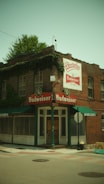 A corner building with red brick walls prominently displaying Budweiser signage and Sundecker's branding. The ground floor appears to be a closed bar or restaurant with green awnings and large windows. There is an old street lamp and a stop sign next to the building. The upper part is covered with green ivy, and there are several windows. The street is empty and the sky is a muted greenish hue.