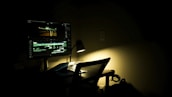 A dimly lit workspace with a computer monitor displaying a video editing software. The room is dark, with a single desk lamp providing focused lighting on the desk. A chair is positioned in front of the desk, and some cables are visible on the floor.