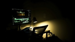 A dimly lit workspace with a computer monitor displaying a video editing software. The room is dark, with a single desk lamp providing focused lighting on the desk. A chair is positioned in front of the desk, and some cables are visible on the floor.