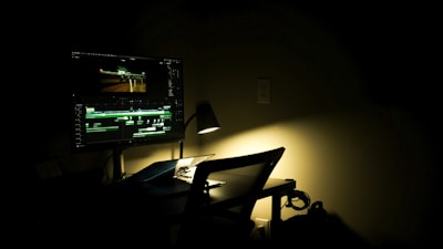 A dimly lit workspace with a computer monitor displaying a video editing software. The room is dark, with a single desk lamp providing focused lighting on the desk. A chair is positioned in front of the desk, and some cables are visible on the floor.