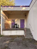 The photo shows a small loading dock area with two purple doors, one with a small window. Three trash bins and several wooden pallets are placed against the walls. A short flight of concrete stairs leads up to the platform, which is surrounded by an industrial-style railing. The area is lit by two ceiling lamps, and dappled sunlight creates patterns on the walls.