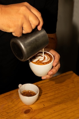 A close-up view of a person pouring milk from a jug into a cup of coffee, creating intricate latte art on the surface. The setting appears cozy with a wooden table and another small cup of coffee in the background.