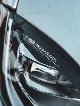 A close-up view of a car's headlight featuring 'JAGUAR LED TECHNOLOGY' branding. The image highlights the sleek and modern design of the headlight, showcasing materials like glass and metal.