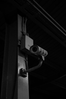 A surveillance camera is mounted on a metal pole, extending from a bracket. The setting appears to be industrial or urban, with shadows creating a dark, moody atmosphere.