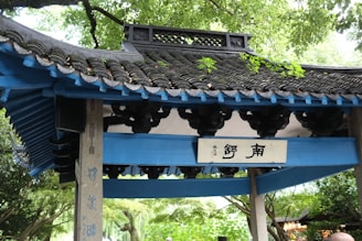 A traditional Asian architectural structure with a blue tiled roof supported by wooden beams. The structure features intricate carvings and a sign with Chinese characters. Surrounding the structure are lush green trees, creating a serene and peaceful environment.