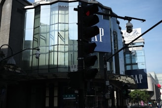 A modern building with large glass windows and two signs that say GAP. There is a traffic light in the foreground with a red light illuminated. The building reflects parts of the street and other structures. Additional elements include streetlights and security cameras.