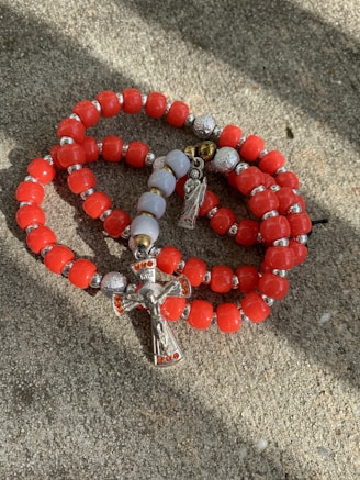 A set of red and silver prayer beads with a crucifix and a small angel charm are laid on a textured stone surface. The beads are predominantly red with interspersed metallic silver and white beads.