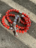 A set of red and silver prayer beads with a crucifix and a small angel charm are laid on a textured stone surface. The beads are predominantly red with interspersed metallic silver and white beads.